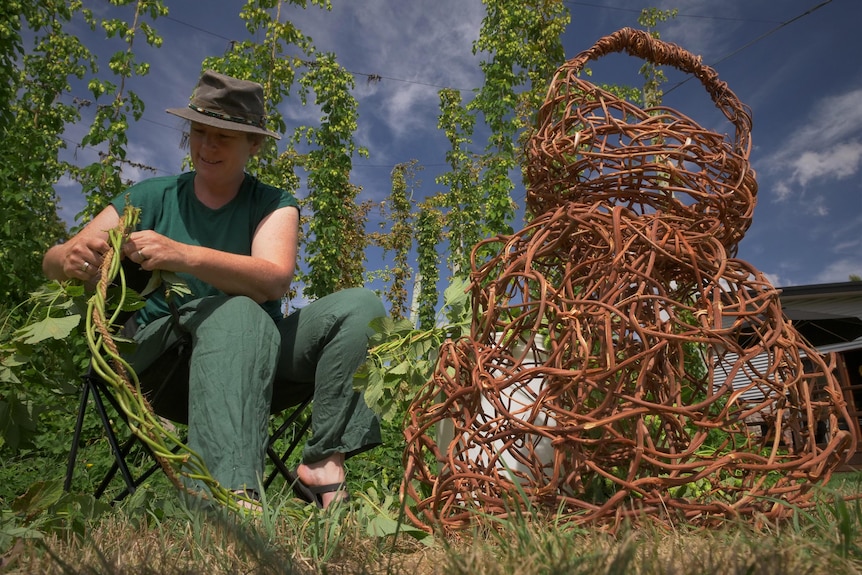 A seated woman wearing a hat, twisting some green vines, with some woven creations sitting beside her.