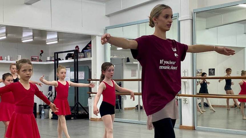 An adult ballerina leads a class of young ballerinas in red and black leotards in a dance studio with mirrored walls.