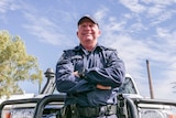 A police officer leans on the front of a police car, arms folded, looking out above the camera.