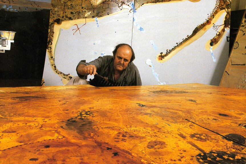 A man leans over looking at large art canvas of browns and oranges