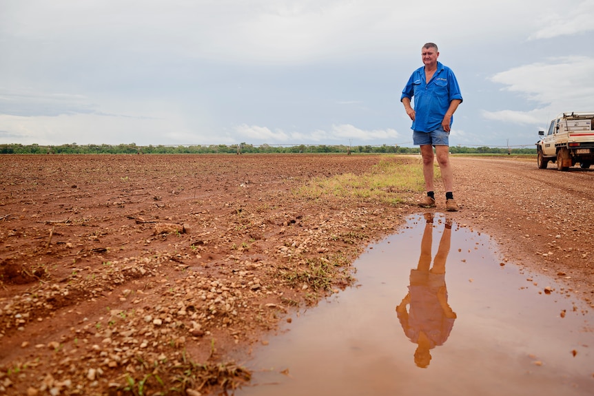 A man in a blue workshirt stands in front of a puddle on a farm in the Top End.