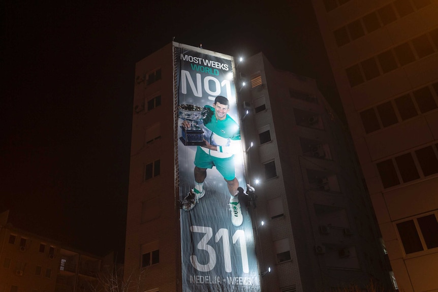 A spotlight shines on a billboard on a tall building, celebrating tennis player Novak Djokovic.