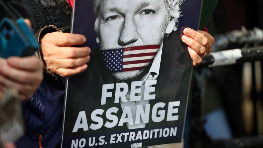 Two hands holding a poster with a picture of a man on it with his mouth covered and the words "Free Assange"
