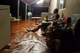 A man sits on a verandah of a building with brown floodwater raging around at his ankles.