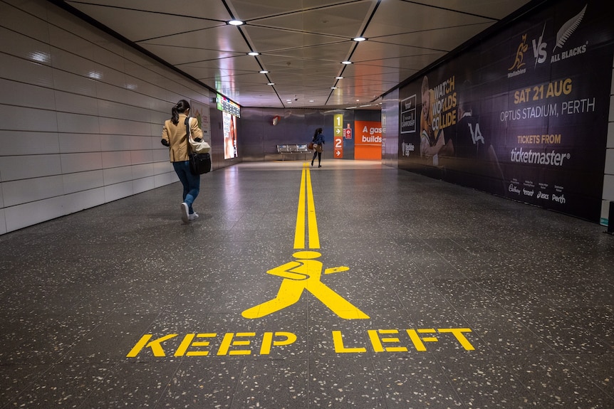 Two people walk through an underpass at Perth train station with 'keep left' written in yellow on the ground.