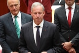 Richard Colbeck, wearing a dark suit with a red pin on his lapel, being sworn into Federal Parliament.