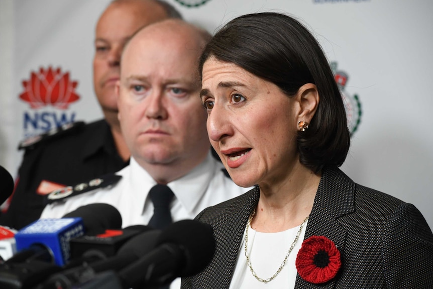 woman speaking at a press conference, serious looking man behind her