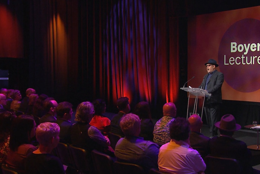 A man in a black suit standing at a lectern in front of a crowd