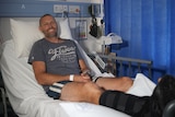 A smiling man lays on a hospital bed with a moon boot on his right leg and foot.