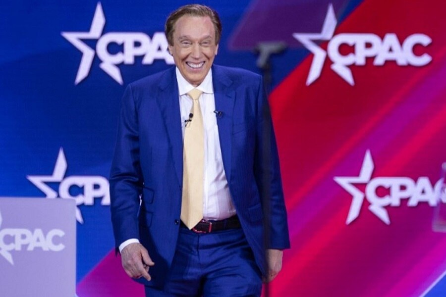 An older man with dyed hair in a blue suit walks onto the stage at CPAC.