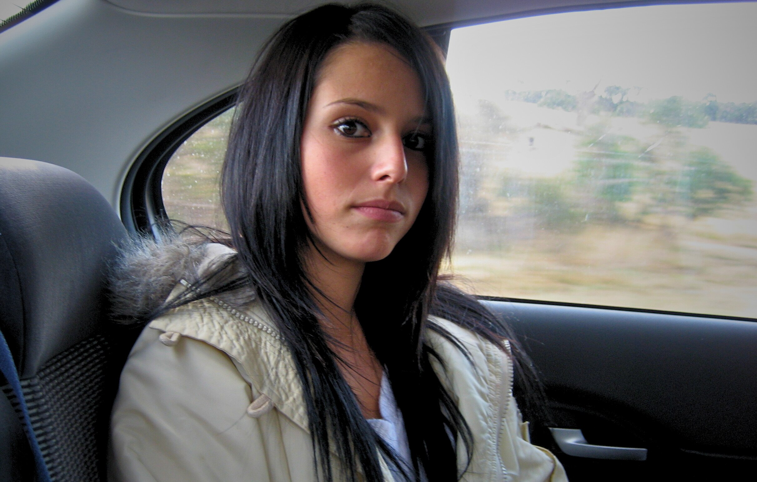 A young woman with long dark hair wears a cream jacket sitting in a car