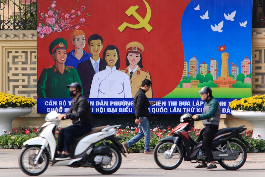 Un hombre camina por la calle mirando su teléfono entre dos personas en motocicleta, con una obra de arte comunista de fondo.