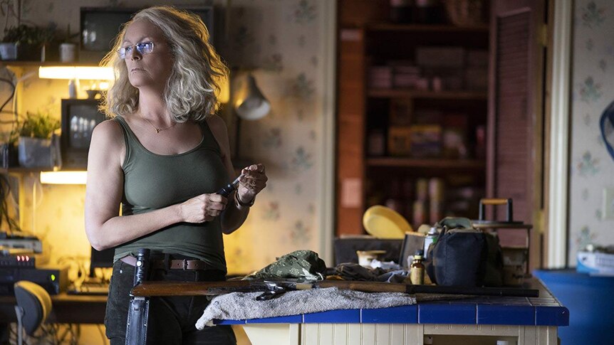 Jamie Lee Curtis stands by a table with a gun on it