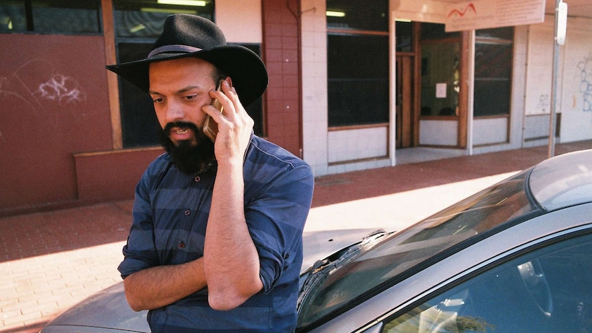 A man with a black beard and black, broad-brimmed hat speaks on a mobile phone.