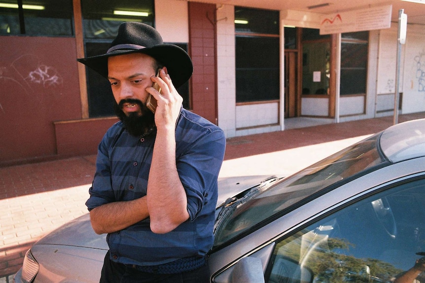 A man with a black beard and black, broad-brimmed hat speaks on a mobile phone.