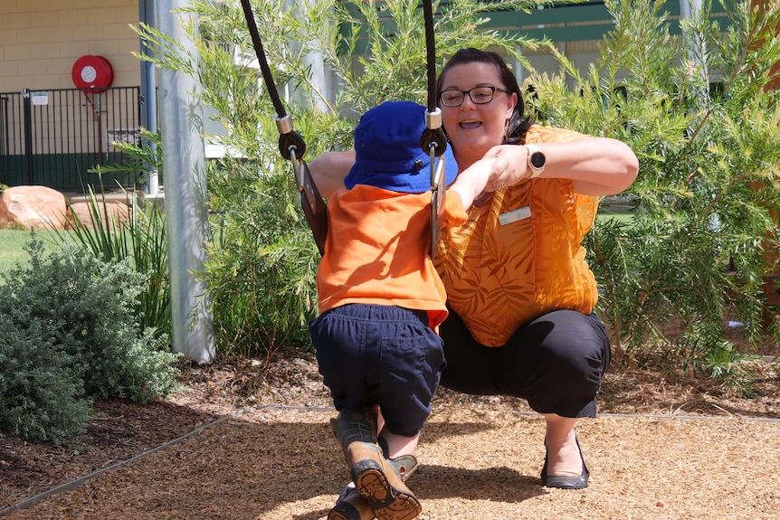 woman in orange shirt pushes child on swing at playground