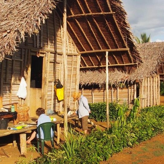 Two women outside a wooden hut on a sunny day, one sitting on a green plastic chair while working, the other standing