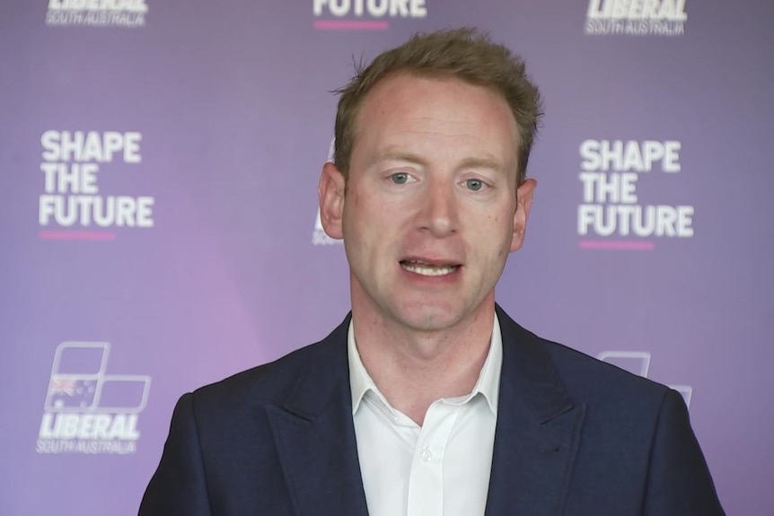 A man with light brown hair wearing a blue blazer in front of a Liberal banner