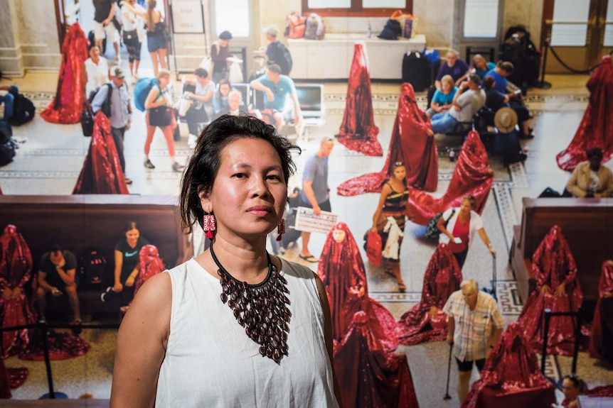 An asian woman in a crisp white dress and big jewelry stands in front of a photo of her artwork