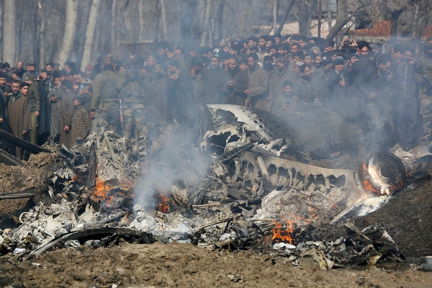 A large crowd gathers around the smoking wreckage of a downed aircraft