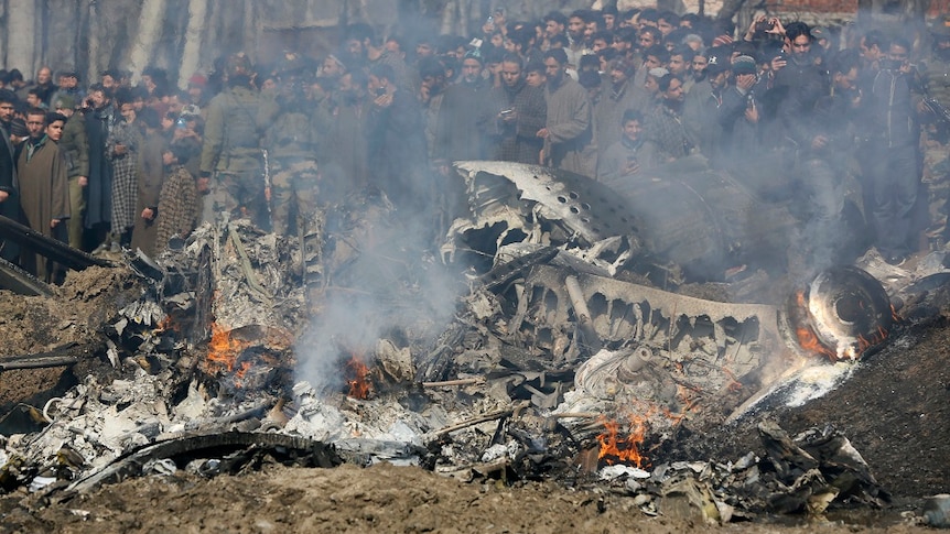 A large crowd gathers around the smoking wreckage of a downed aircraft