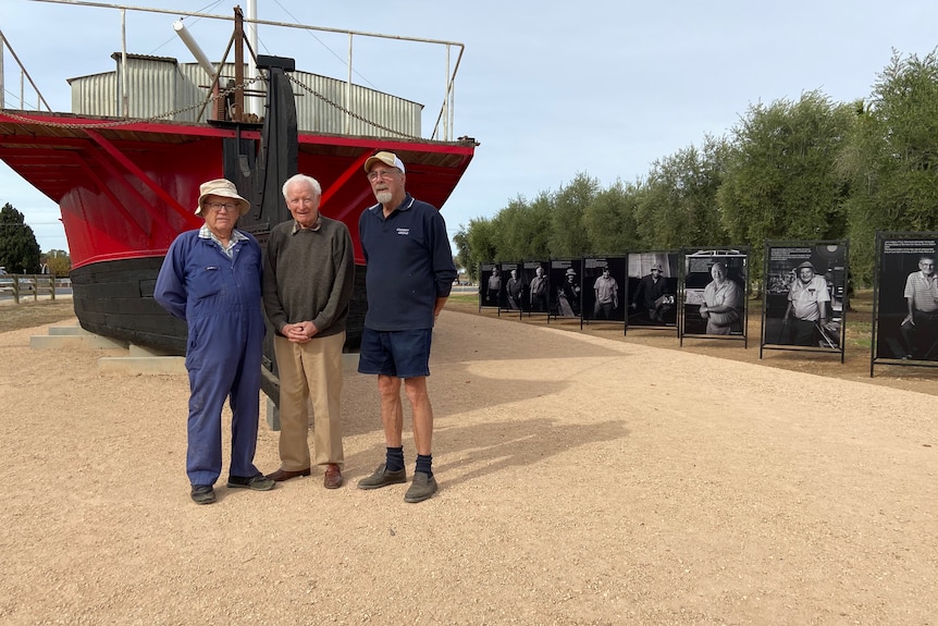 Three men stand in front of a large red and black barge.