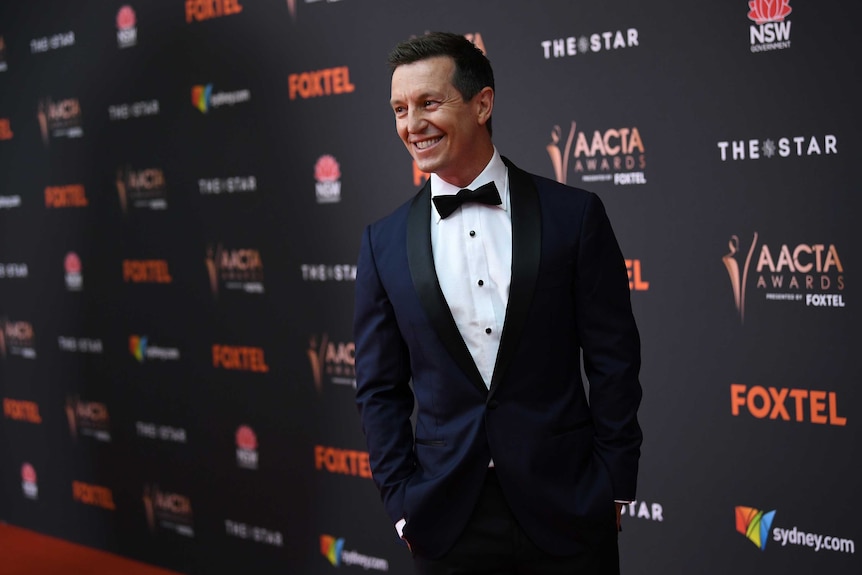 A smiling man wears a navy tuxedo on the red carpet.
