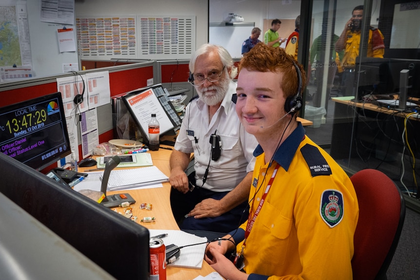 A young man sitting in front of an older man at the communications desk in an emergency  incident control centre.