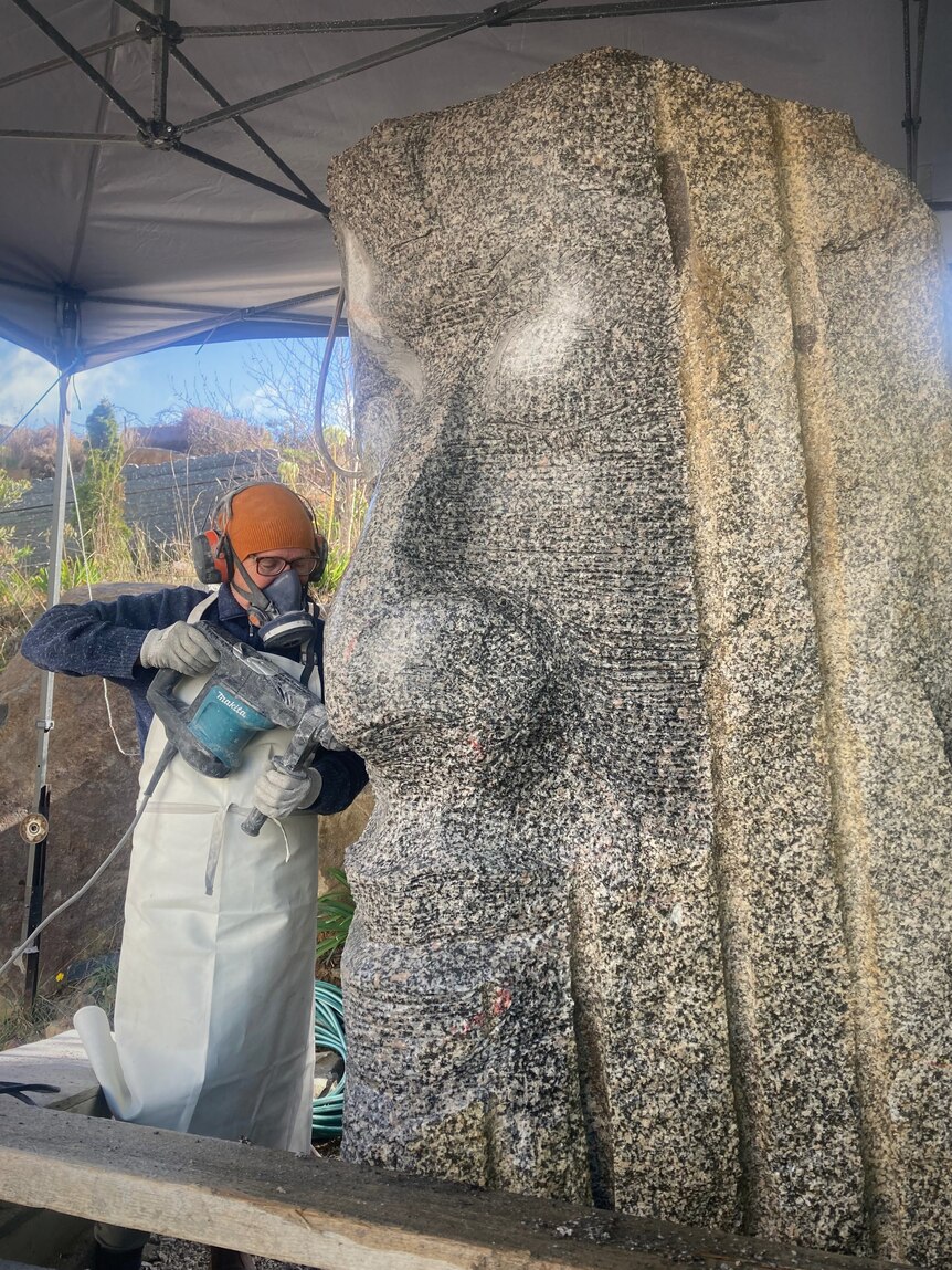 A woman using a mini jack hammer to carve a very large granite sculpture