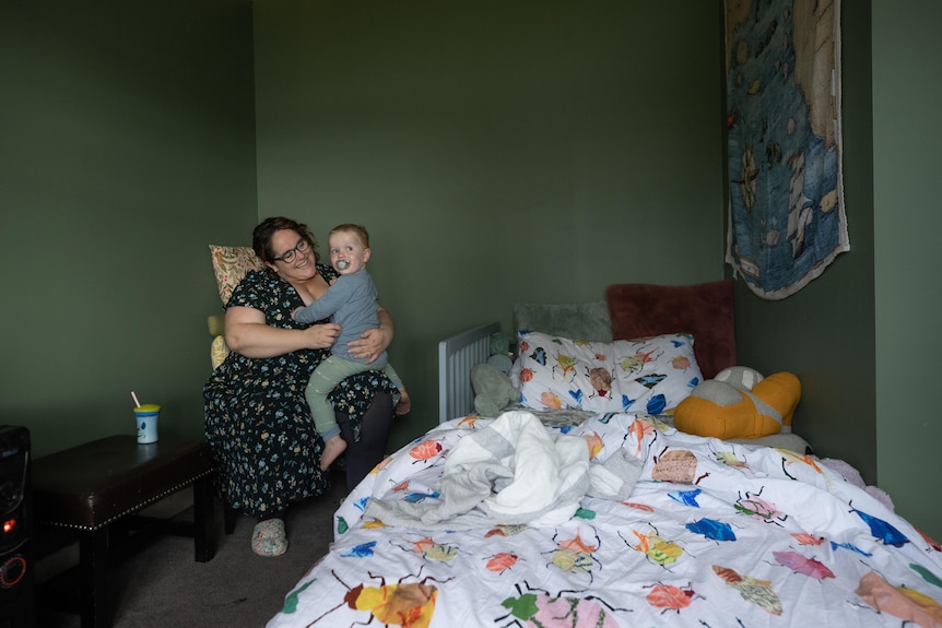 Emma Helks sits on a chair holding 2-year-old Theo next to his bed, which is covered with a bright doona