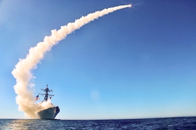 A smoke trail curves into the horizon from a large naval destroyer at sea.