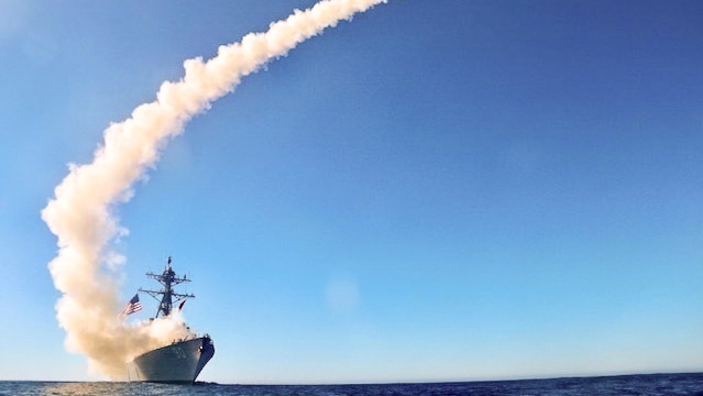 A smoke trail curves into the horizon from a large naval destroyer at sea.