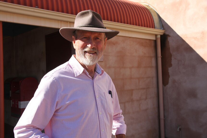 An older man with a neat, white beard stands in the sun wearing a broad-brimmed hat.