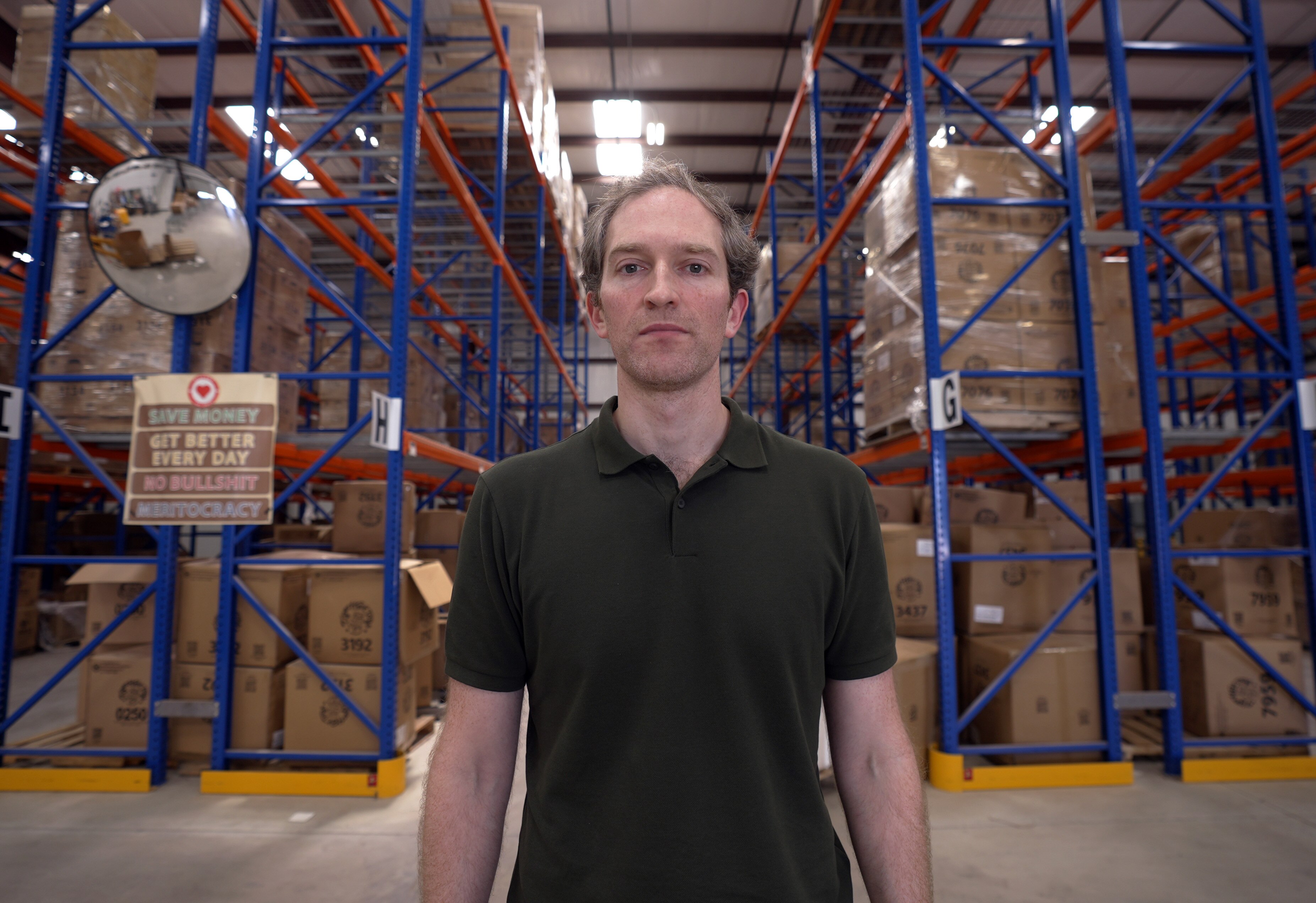 A man stands looking at the camera. Behind him is the interior of a large warehouse.