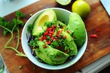 A bowl filled with avocado and garnishes sits on a chopping board.