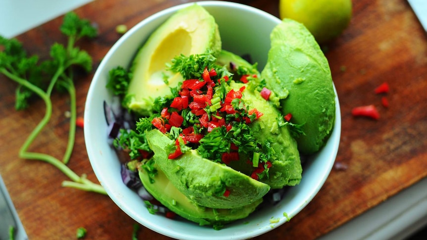 A bowl filled with avocado and garnishes sits on a chopping board.