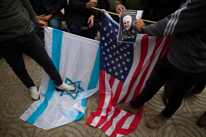 You look down on a crowd of bodies stepping on the Israeli and American flag while a hand holds the photo of Qassem Soleimani.