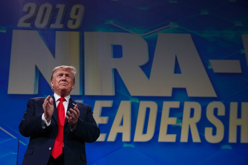 Donald Trump stands in front of a NRA banner about to clap his hands