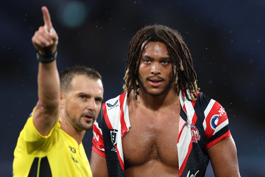A Sydney Roosters player with his shirt ripped open stands on the field as the referee points him to the sheds after a send-off.