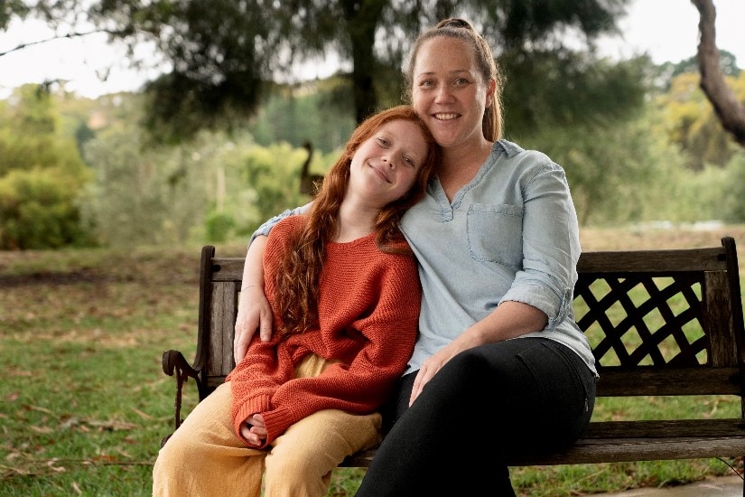 A young girl wearing an orange dress sits next to her mother on a park bench. 