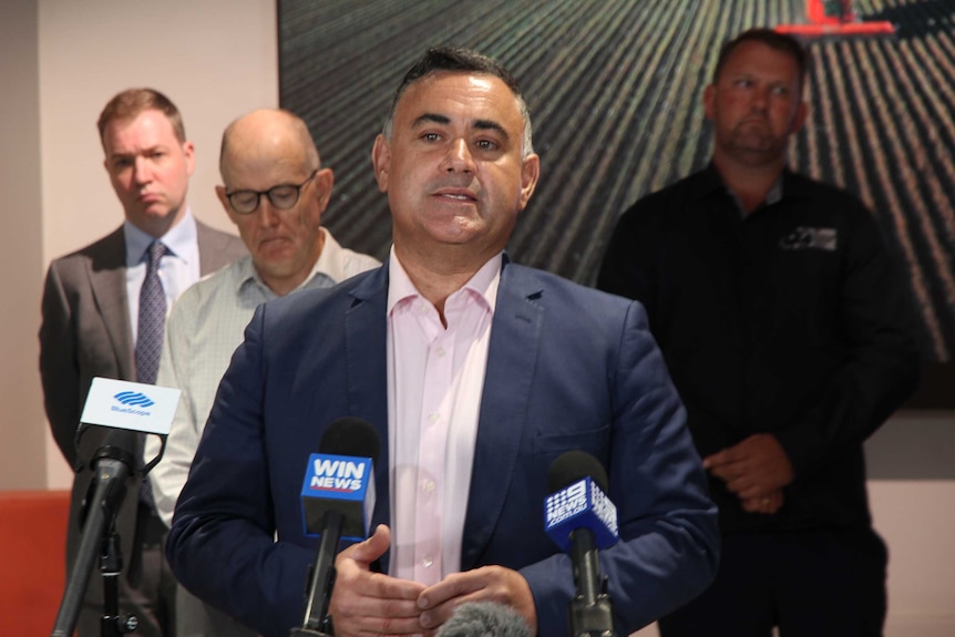 A man stans in front of microphones talking to the media with three men standing behind him.