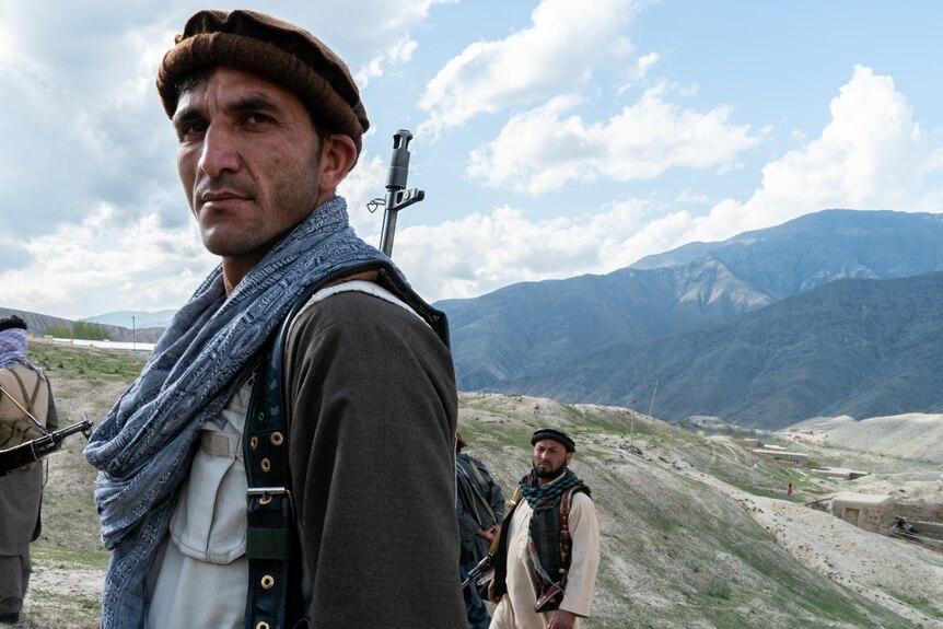 A man with a gun on his shoulder stands side-on to the camera with mountains and valleys behind him