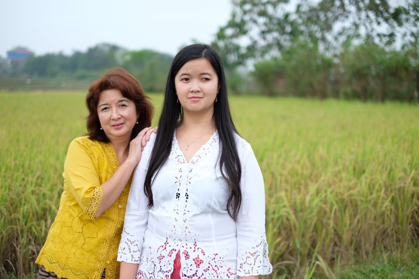 Dua orang perempuan berdiri berdampingan dengan latar belakang sawah.