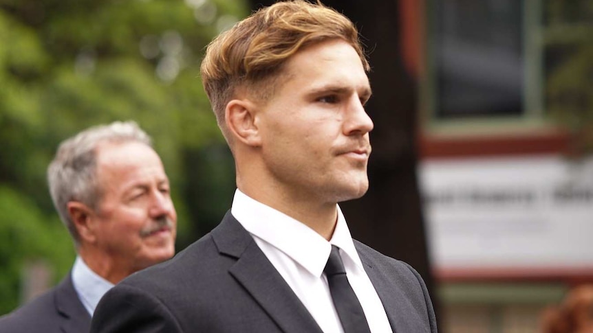 A man with short, fair hair, wearing a suit, stands on a street.