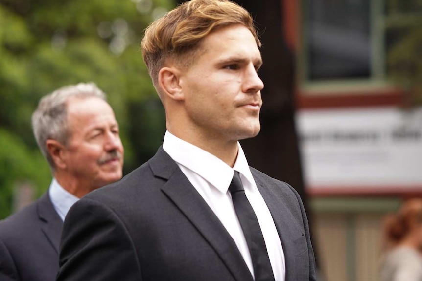 A man with short, fair hair, wearing a suit, stands on a street.