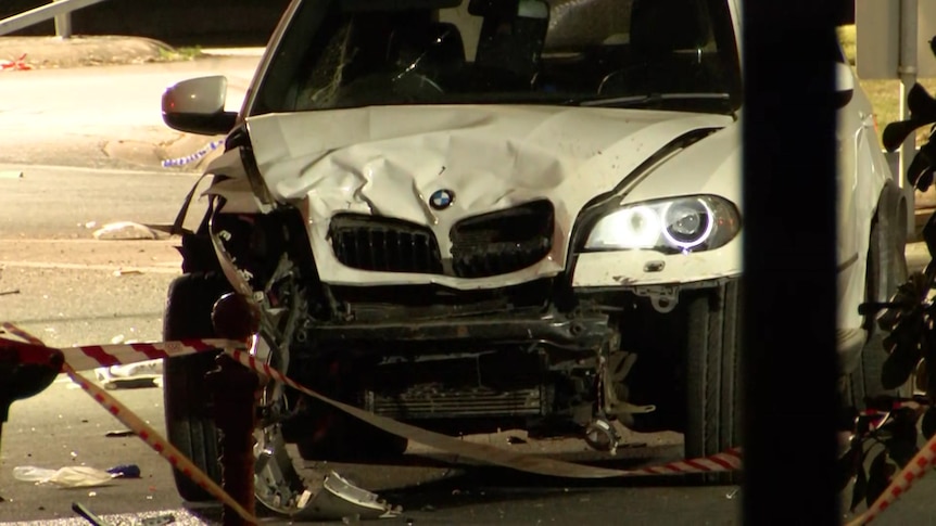 The crumpled front of a white BMW SUV, surrounded by emergency services tape.