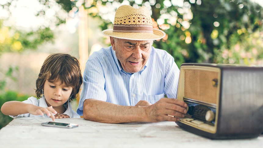 A grandfather and young grandchild listen to music, one on an old radio, the other on a mobile phone.