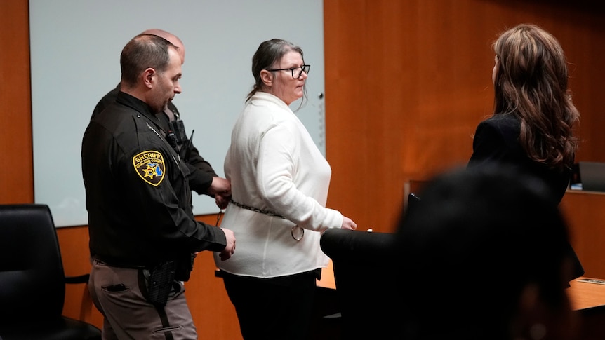 Jennifer Crumbley is escorted out of court, wearing white jumper and black pants. Two police by her side.