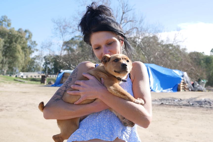 A woman with her hair in a bun holds a small puppy to her chest while standing in front of makeshift tents.