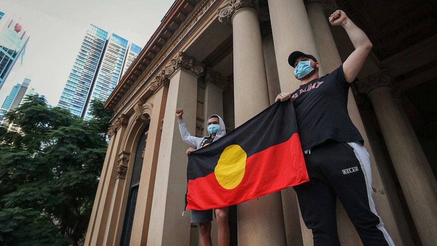 Two men carrying an Aboriginal flag raise their clenched fists.